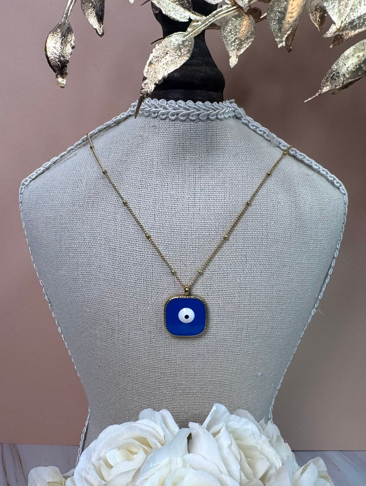 Necklace with a blue evil eye pendant on a mannequin bust with white flowers and gold leaves.